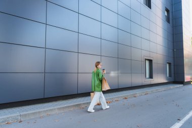 Alışveriş torbaları ve paket içkileriyle genç şık bir kadın gri duvarda yürüyor.