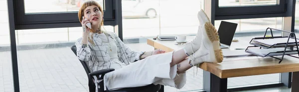 trendy tattooed businesswoman talking on smartphone while sitting with legs on desk in office, banner