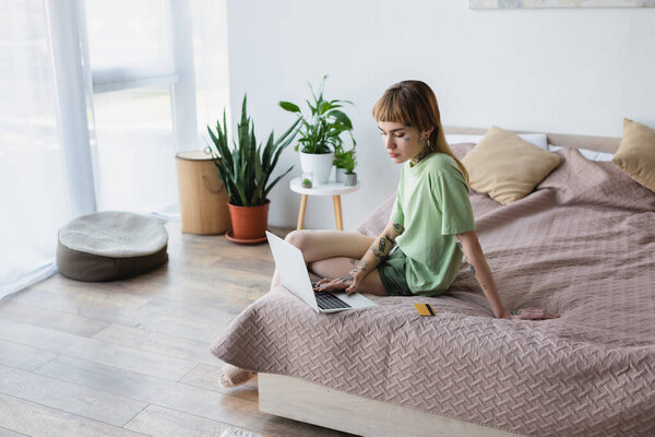 женщина с татуированным телом печатает на ноутбуке, сидя на кровати рядом с кредитной картой