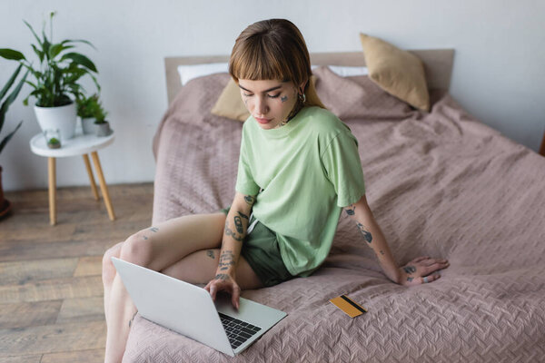 молодая татуированная женщина с помощью ноутбука, сидя на кровати рядом с кредитной картой