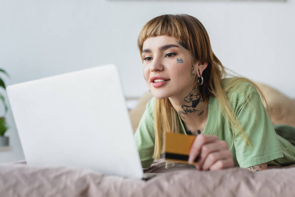 улыбающаяся татуированная женщина с кредиткой в руках, лежащая на кровати рядом с размытым ноутбуком