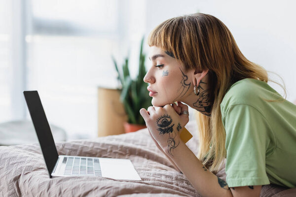 боковой вид татуированной женщины с кредитной картой, смотрящей на ноутбук, лежа на кровати