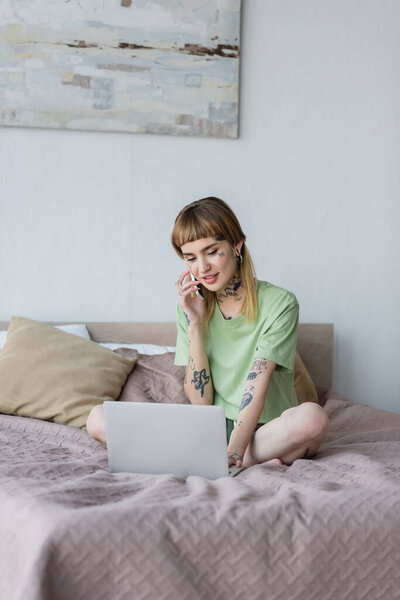 улыбающаяся татуированная женщина, работающая на ноутбуке и разговаривающая по мобильному телефону на кровати дома