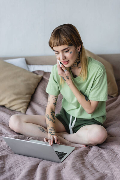 положительная женщина с татуированным телом, печатающим на ноутбуке во время звонка на мобильный телефон в спальне