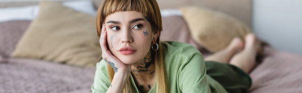 young woman with tattoo and piercing looking away while lying on bed, banner