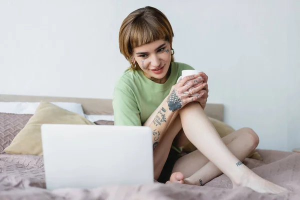 tattooed woman with cup of tea smiling while watching movie on blurred laptop in bedroom