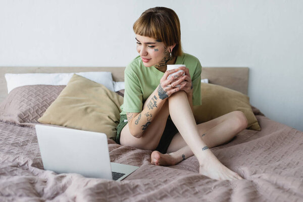 татуированная босиком женщина улыбается сидя на кровати с чашкой чая и смотря фильм на ноутбуке