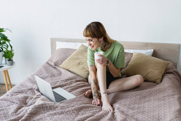 молодая татуированная женщина сидит на кровати и смотрит кино на ноутбуке