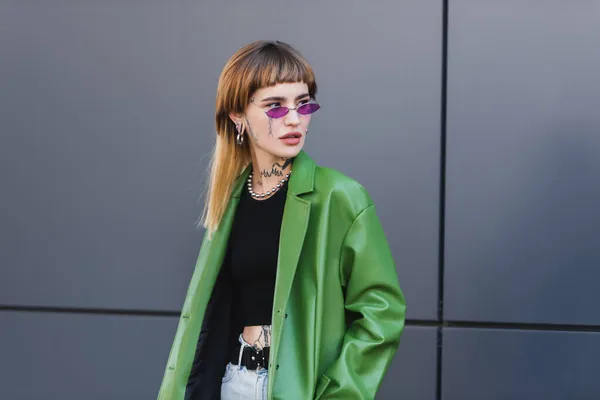 tattooed woman in green leather jacket and trendy eyeglasses looking away on city street