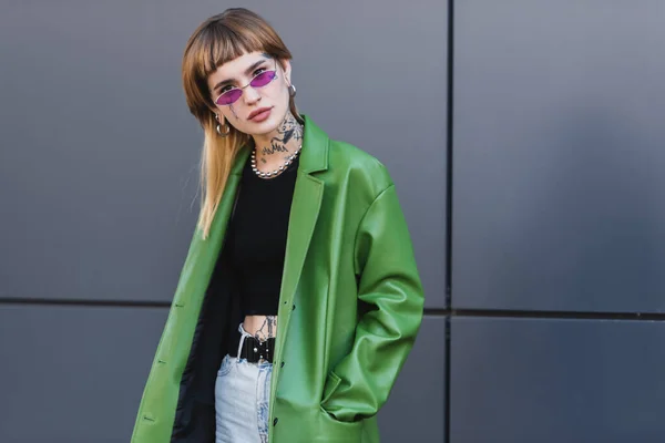 tattooed woman with piercing looking at camera while posing with hand in pocket of green jacket