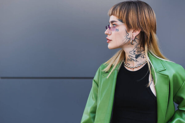 tattooed woman in stylish eyeglasses, green jacket and silver necklace looking away near grey wall