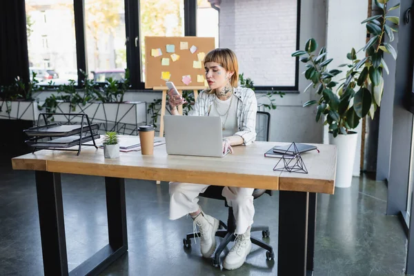 full length view of tattooed businesswoman working with smartphone and laptop in modern office