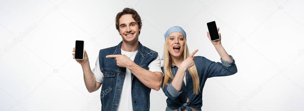 cheerful couple in denim clothes pointing at smartphones isolated on white, banner