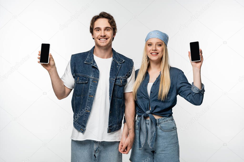 stylish and cheerful couple holding hands while showing smartphones isolated on white