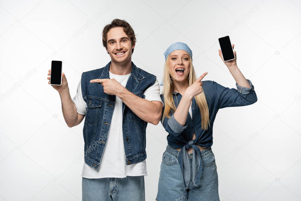 happy stylish couple pointing at mobile phones with blank screen isolated on white