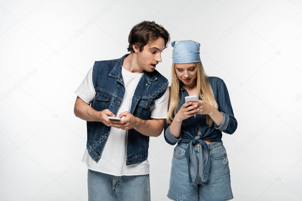 amazed man in denim clothing looking at woman using cellphone isolated on white