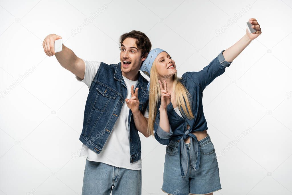 happy couple in denim clothing showing victory sings while taking selfie isolated on white