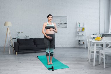 Hamile bir kadın, oturma odasında meditasyon yaparken karnına sarılıyor.