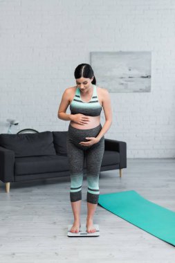 Spor kıyafetli hamile bir kadın, yer ölçeğinde ağırlığını ölçerken göbeğine sarılıyor.