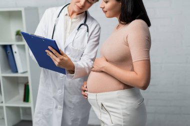 Doktor, muayene odasında hamile bir kadına reçete gösteriyor.
