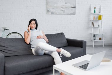 Masadaki bulanık dizüstü bilgisayarın yanında oturan esmer hamile kadın cep telefonundan arıyor.