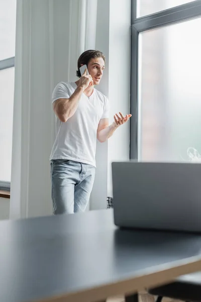 Mladý Muž Gestikuluje Zatímco Mluví Smartphone Blízkosti Rozmazané Notebook Stole — Stock fotografie