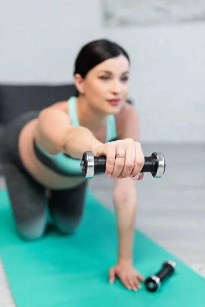 selective focus of dumbbell in hand of blurred pregnant woman training on fitness mat