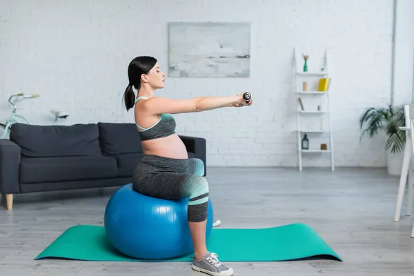 黑发孕妇坐在健身球上用哑铃锻炼的侧视图 — 图库照片