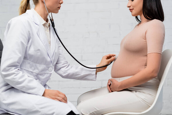 частичный взгляд беременной женщины и врача, осматривающего ее живот стетоскопом во время консультации