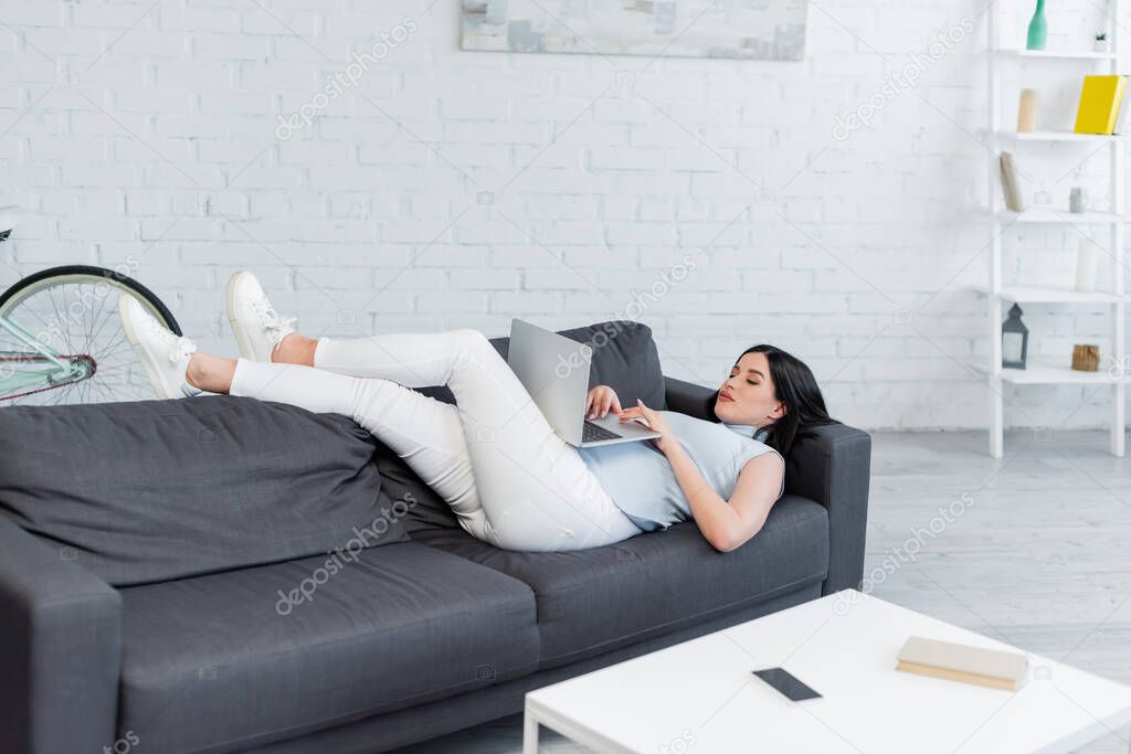 pregnant woman typing on laptop while lying on couch in living room