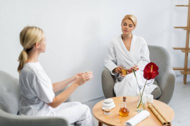 Üzerinde krema kabı olan bulanık spa uzmanına bakarken elinde çay tutan bornozlu bir müşteri.