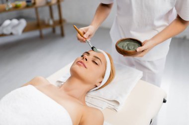 beautician applying clay mask on face of woman lying on massage table clipart
