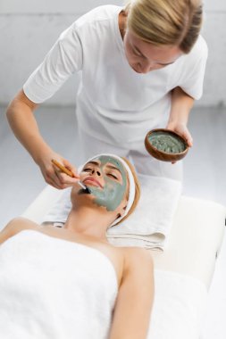 Sarışın güzellik uzmanı masaj masasında yatan kadının yüzüne kilden maske takıyor.