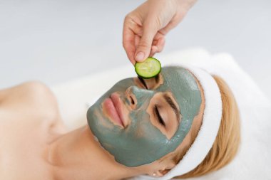 beautician putting cucumber slice on eye of woman in clay mask clipart