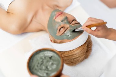 Güzellik uzmanı masaj masasında yatan sarışın kadına kilden maske takıyor.