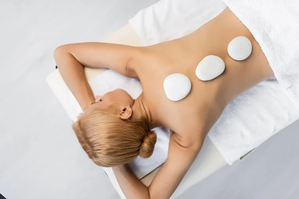 top view of blonde woman with closed eyes receiving hot stone massage in spa center