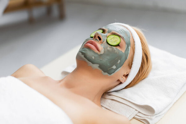 blonde woman with clay mask and cucumber slices on eyes lying on massage table