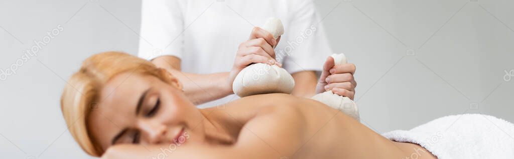 blonde woman with closed eyes receiving massage with herbal bags in spa center, banner