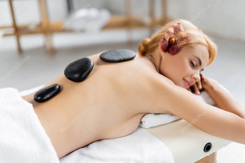 pleased woman with flower in hair receiving hot stone massage in spa center 