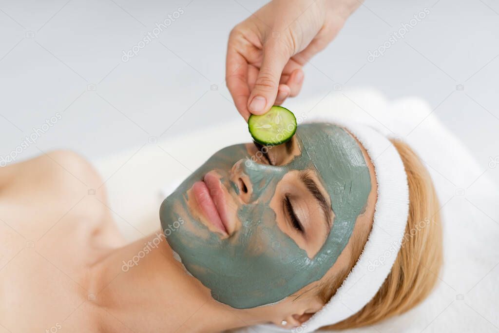 beautician putting cucumber slice on eye of woman in clay mask