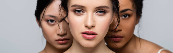 Young woman looking at camera near blurred interracial women isolated on grey, banner 