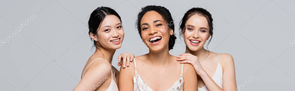 Cheerful multicultural women looking at camera isolated on grey, banner