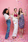 Full length of smiling multiethnic friends with champagne standing under confetti on pink background