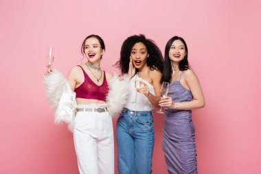 Heyecanlı Afro-Amerikalı kadın pembe renkte izole edilmiş neşeli ırklar arası arkadaşlarının yanında şampanya tutuyor. 