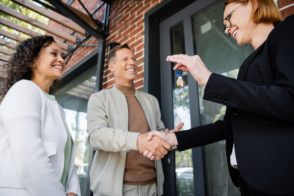 low angle view of cheerful realtor holding key while shaking hands with cheerful man near african american wife