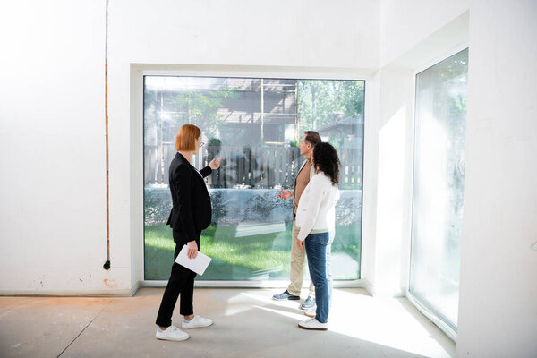 redhead realtor in glasses holding digital tablet while pointing at panoramic window near interracial couple inside of new house