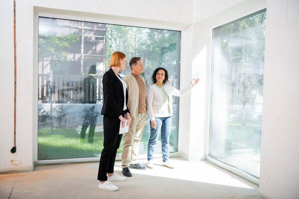 redhead realtor in glasses holding digital tablet near african american woman pointing with hand at panoramic window inside of new house