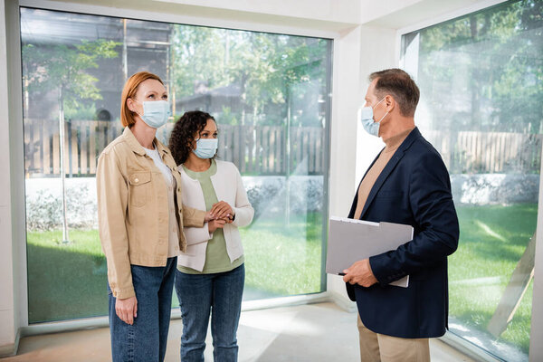 realtor in medical mask holding folder near interracial lesbian couple inside of new house