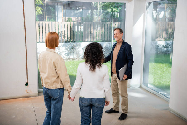 smiling realtor holding folder near interracial lesbian couple inside of new house