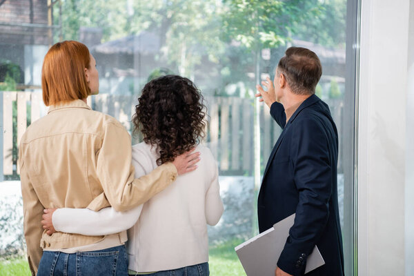 back view of realtor holding folder and pointing at panoramic window near interracial lesbian couple inside of new house